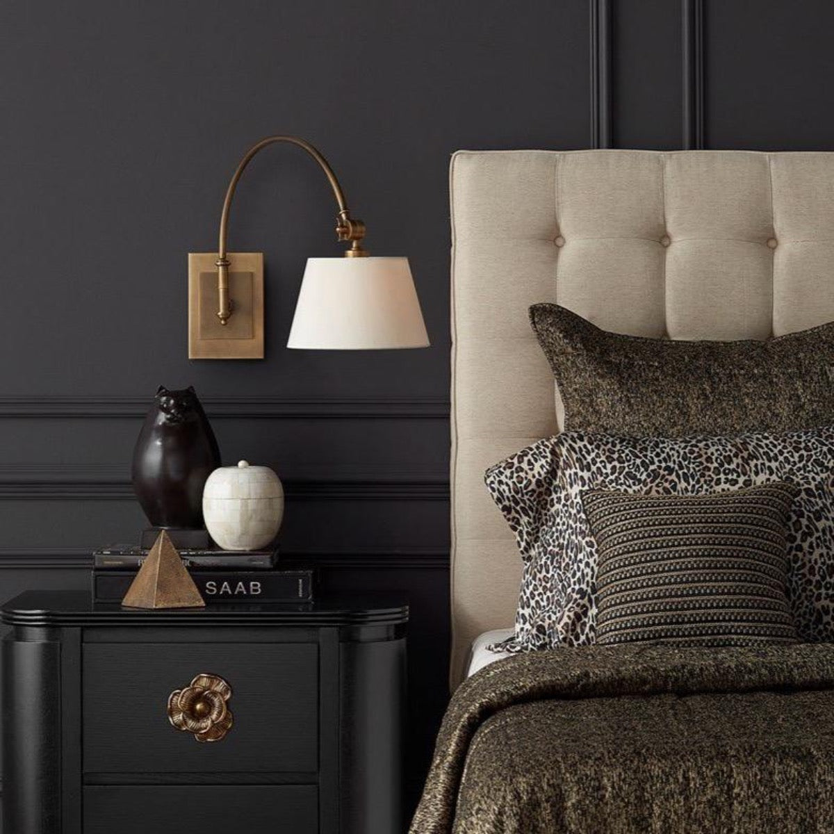 Rennie Swing-Arm Wall Sconce styled in a black wall beside a bed with dark decor. Styled view.