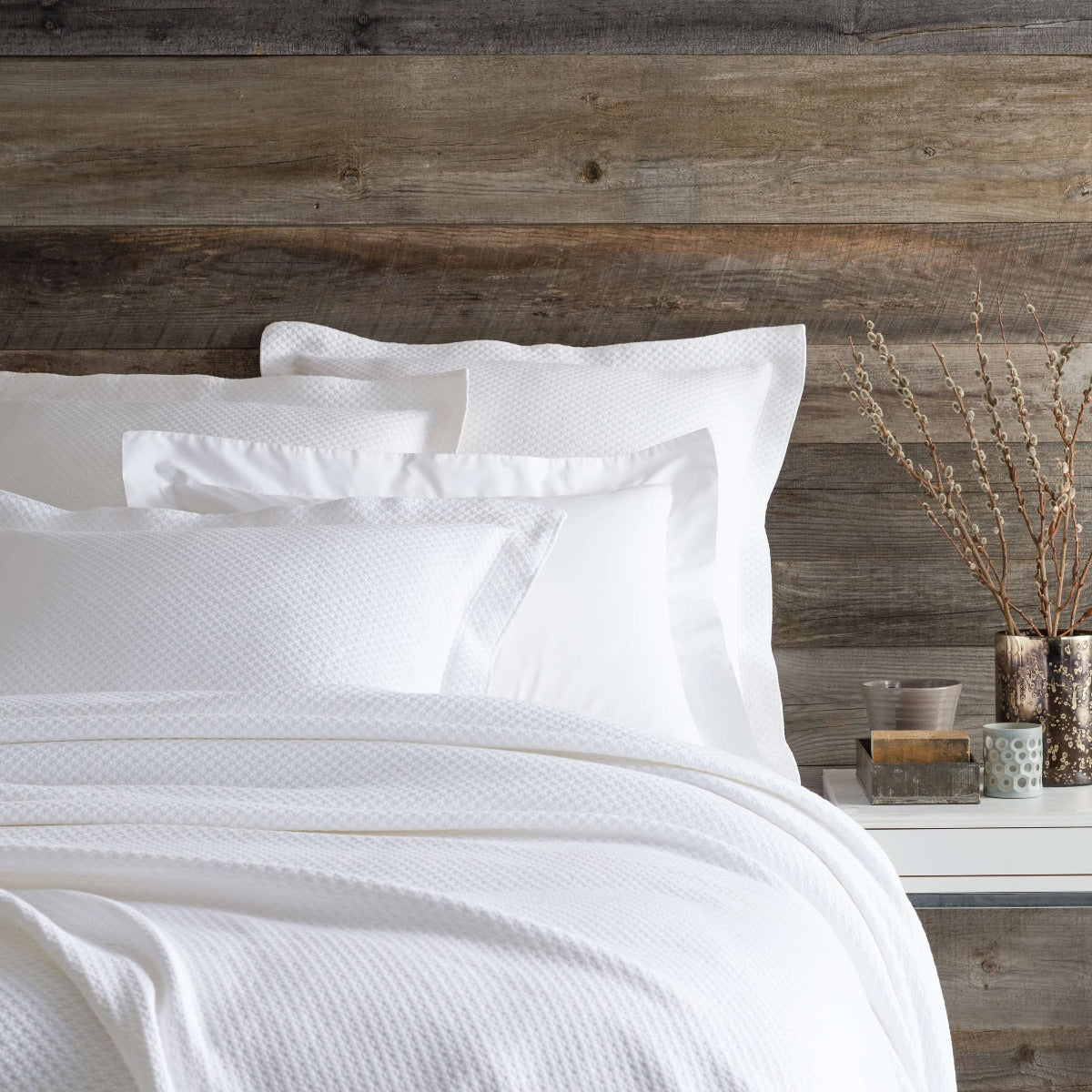 Petite Trellis White Matelasse coverlet styled with white sheets against a wood wall. Styled view. 